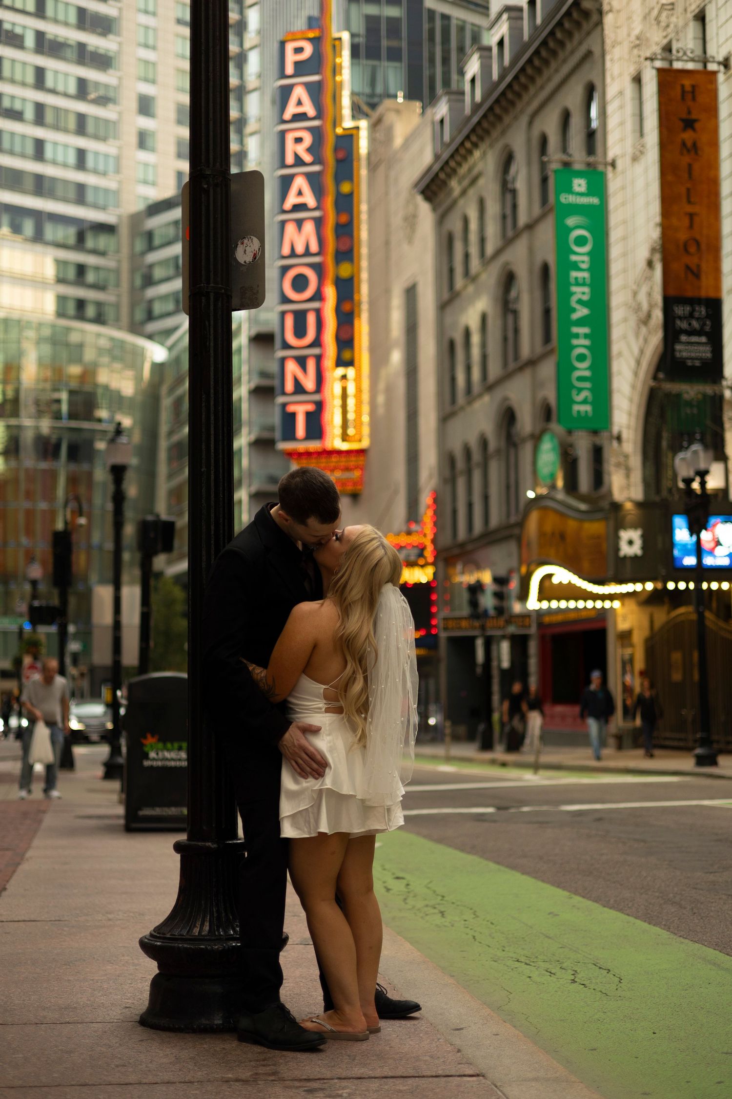 A couple embraces near a lamp post on a city street with neon signs and tall buildings in the background.