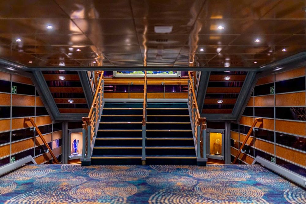 Panoramic view of an empty cruise ship theater with wooden stadium seating and blue carpeted aisles.