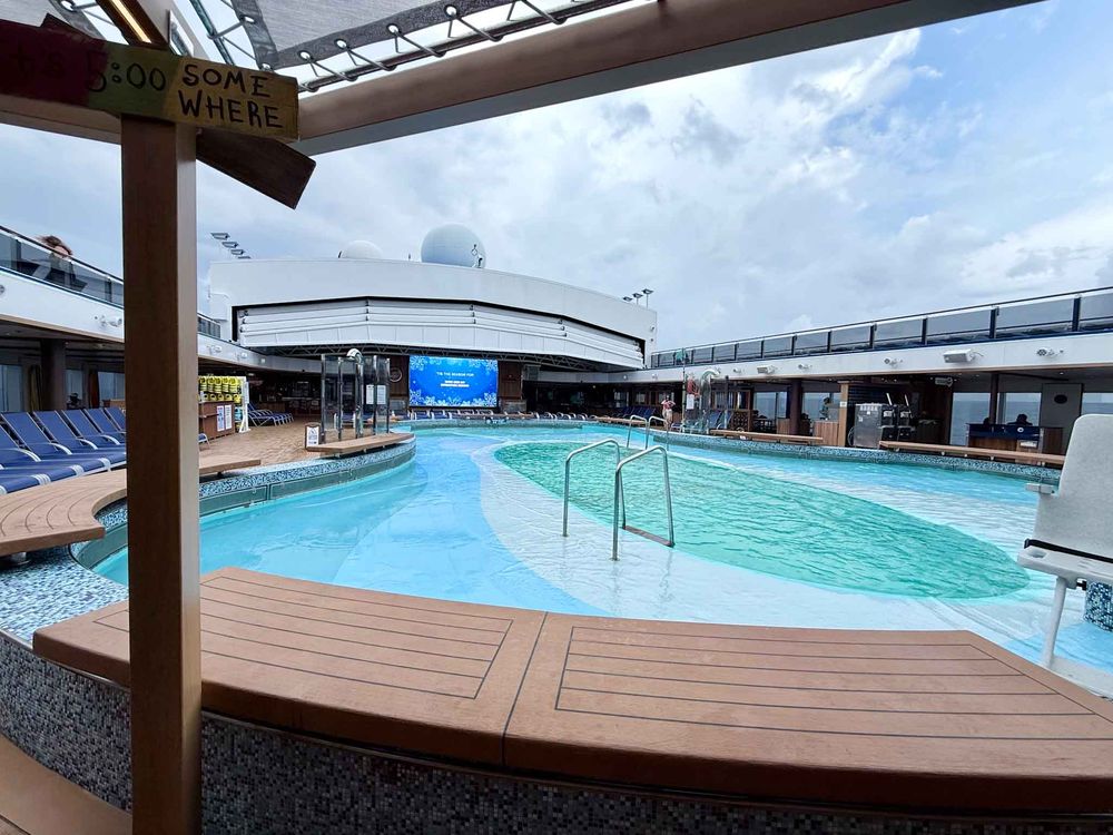 A cruise ship swimming pool deck with lounge chairs and wooden decking under a covered area with blue skies visible.