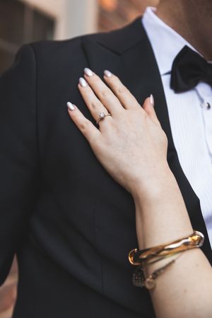 A woman's hand wearing an engagement ring rests on a man's black tuxedo jacket lapel during a formal occasion.