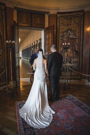 Wedding couple walks down an ornate palace hallway with tapestries and elegant carpeting as their train trails behind.