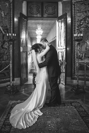 A romantic black and white wedding photo showing a couple embracing in an elegant hallway with chandeliers and ornate decor.