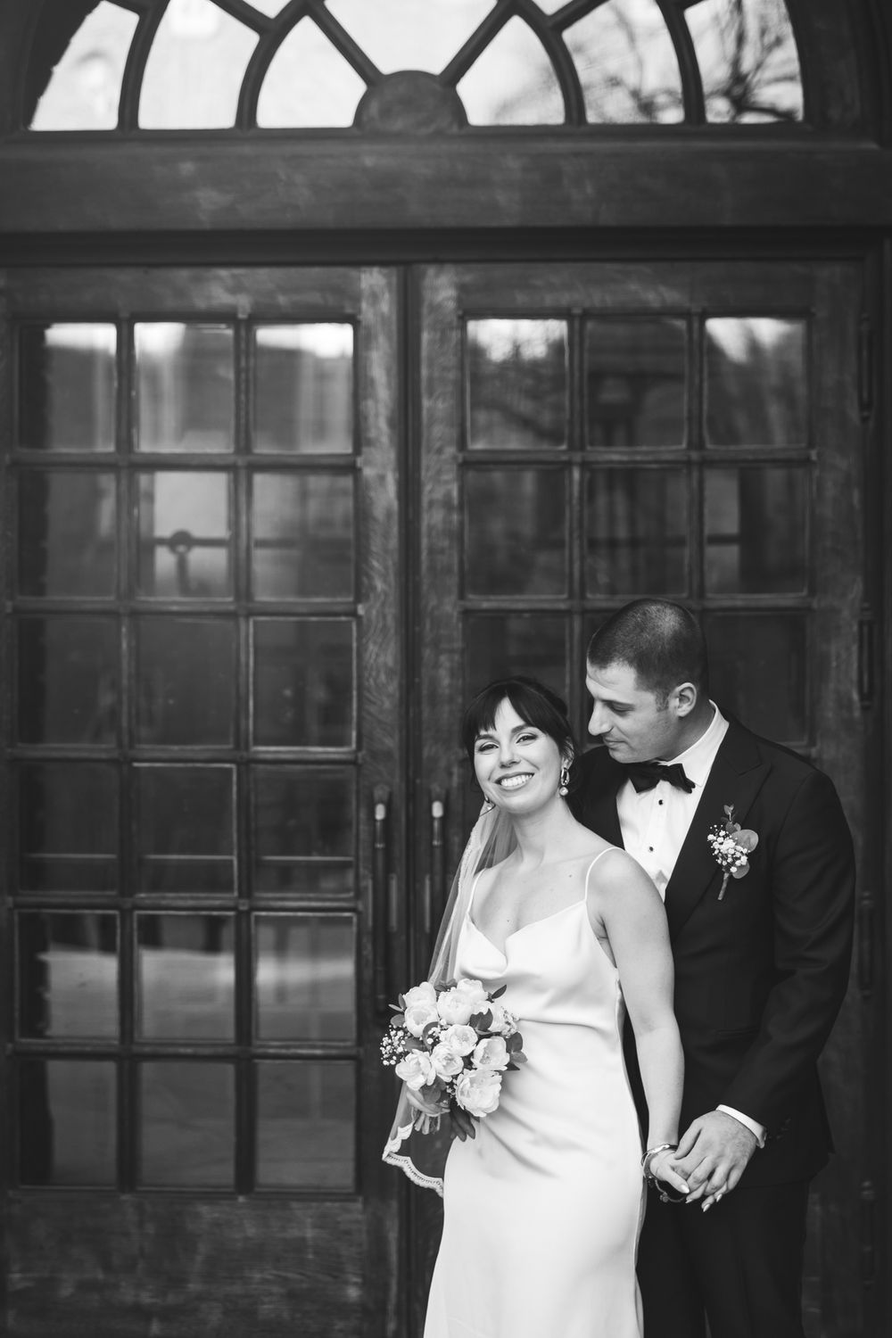 A black and white wedding photo of a bride and groom sharing a joyful moment together outside a building with windows.