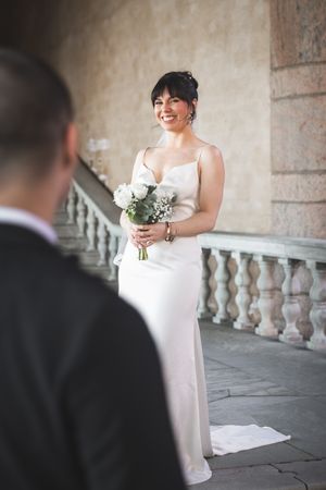 A bride in a white dress holds a bouquet while standing in an elegant stone corridor with decorative balusters.