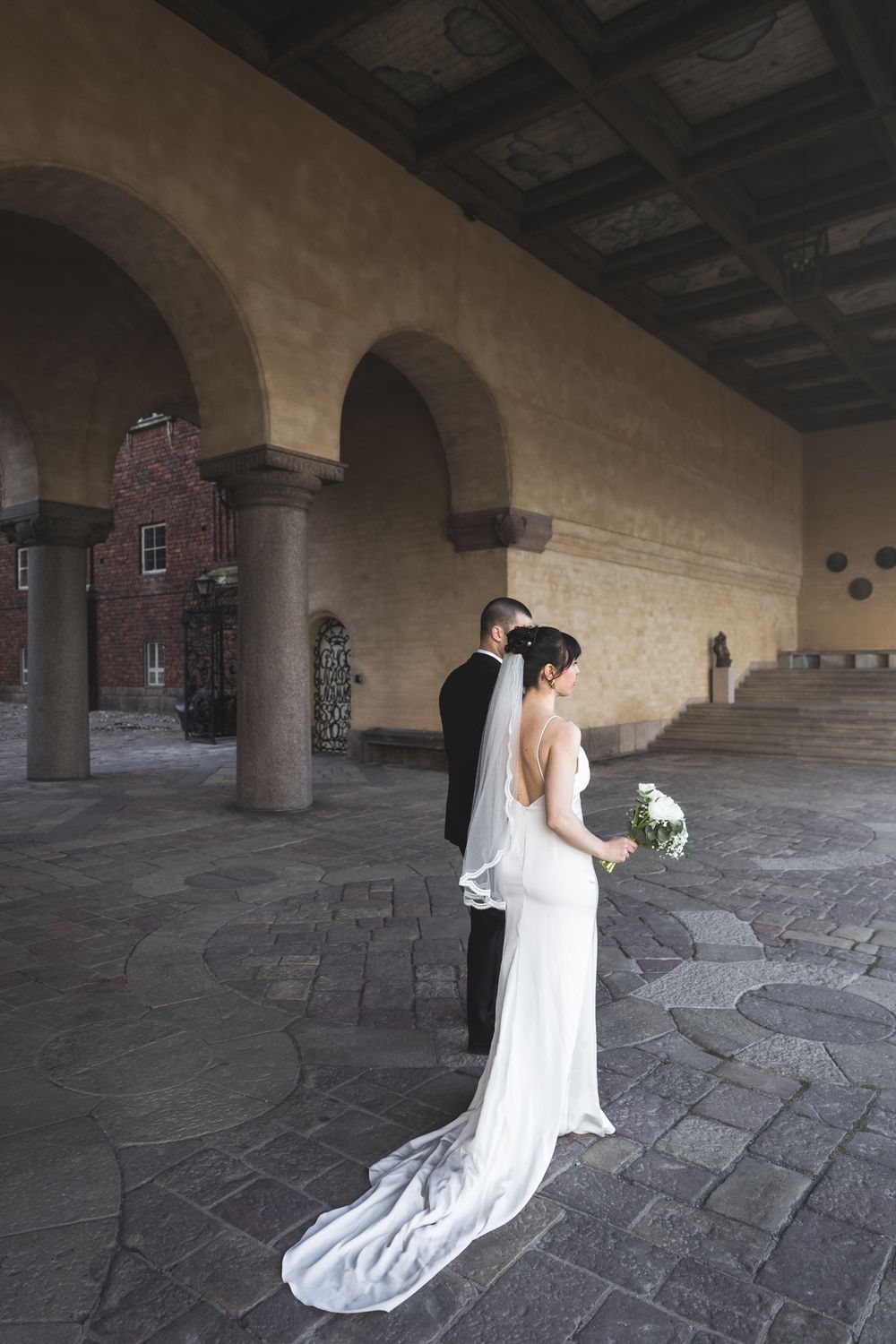 A couple in formal wedding attire embrace under stone archways in a historic courtyard with a long white trailing dress.