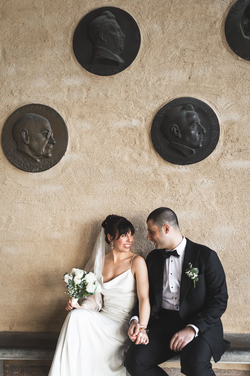A couple sits together on a bench beneath decorative medallions mounted on a textured wall.