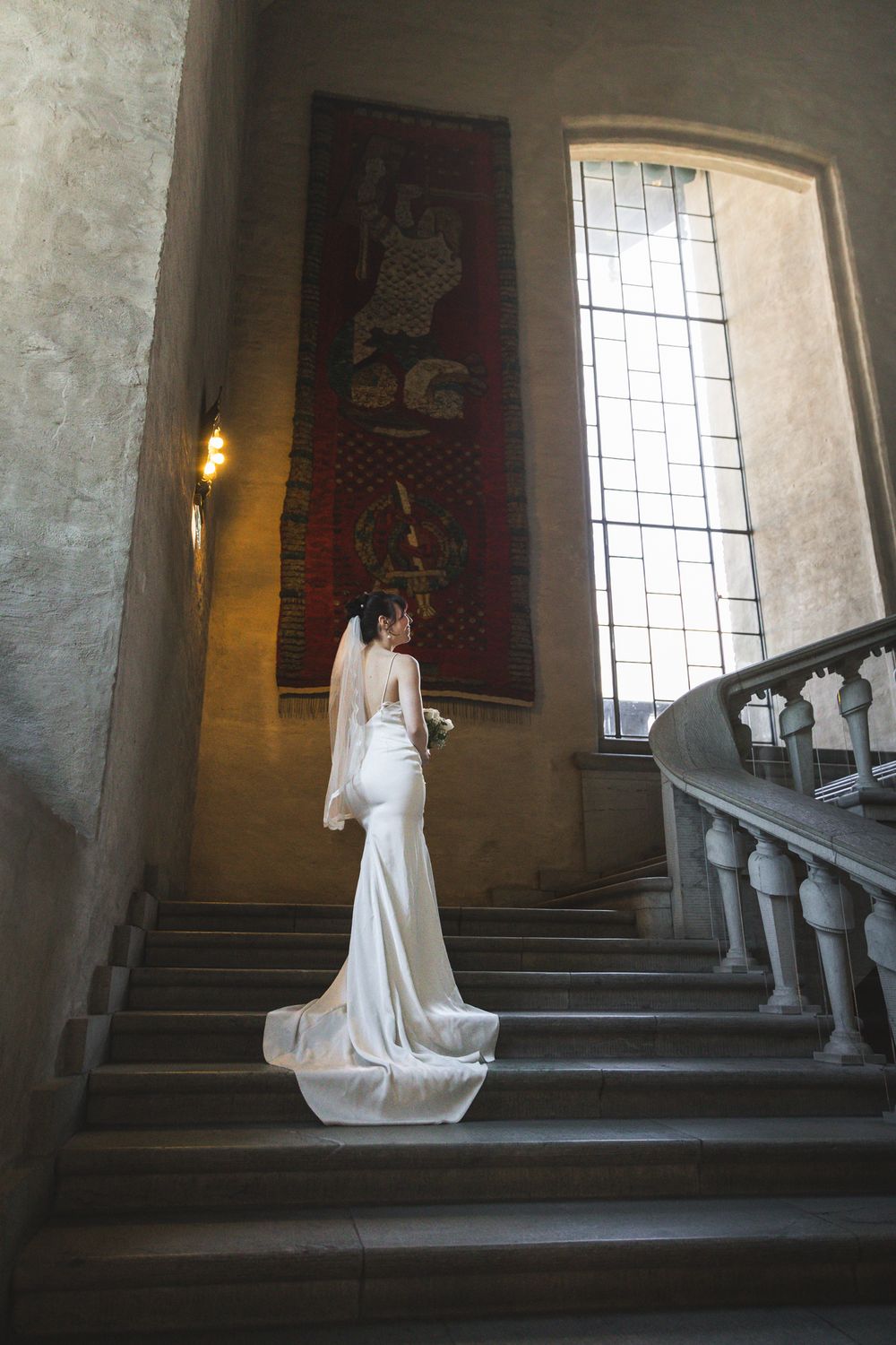 A bride in a flowing white wedding dress poses gracefully on a grand stone staircase in a historic building.