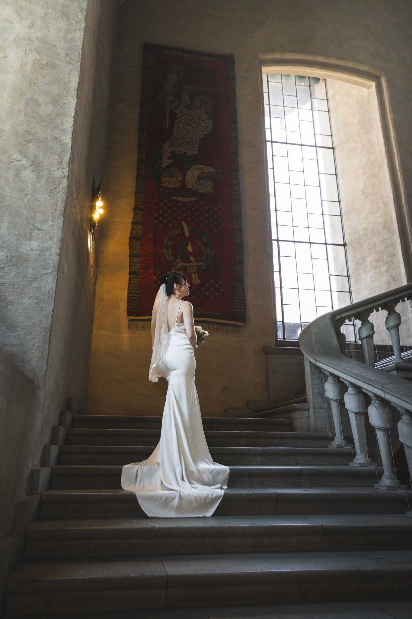A bride in a flowing white wedding dress poses gracefully on a grand stone staircase in a historic building.