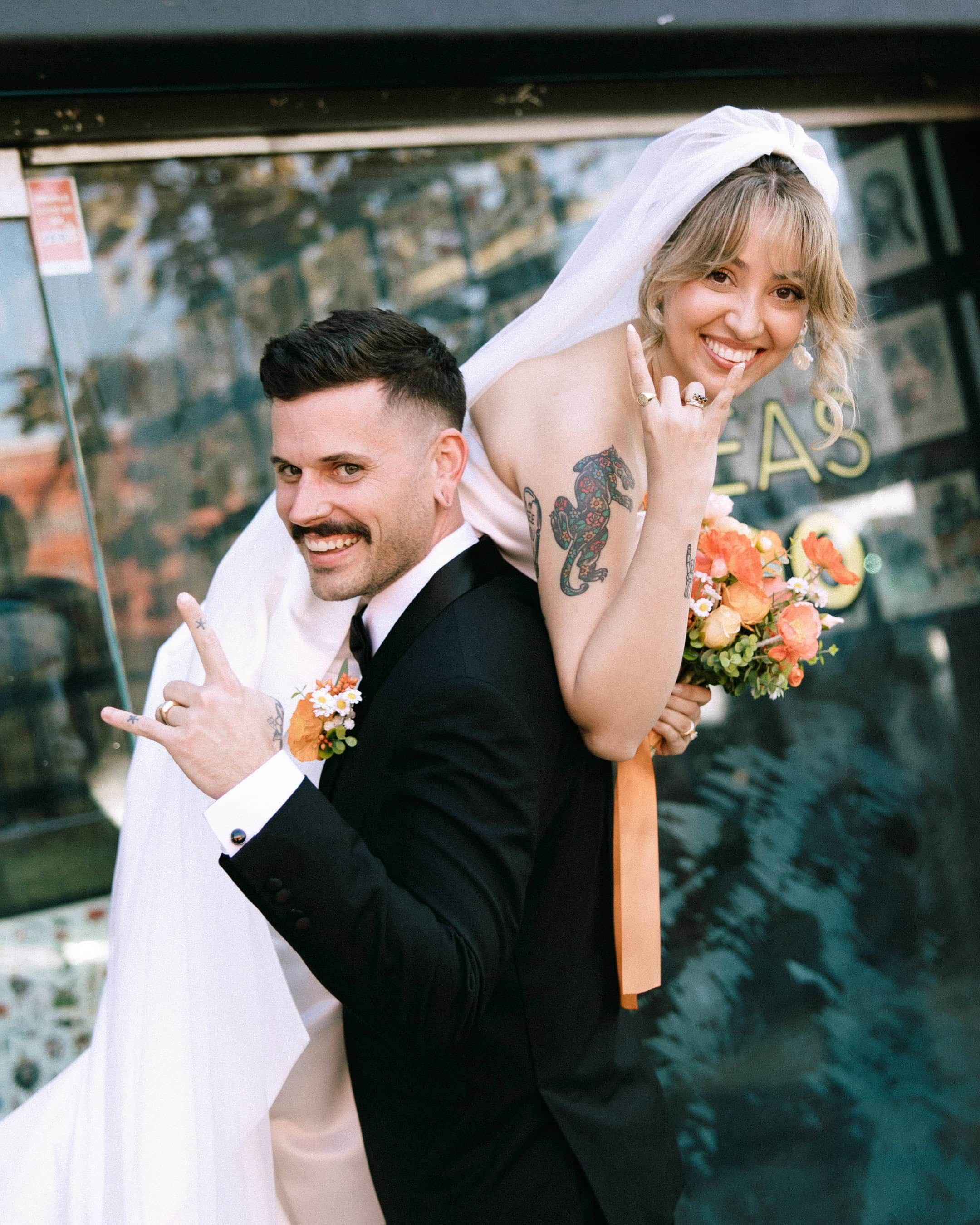 Smiling newlywed couple in wedding attire making rock on hand gestures, bride holding orange bouquet outdoors.