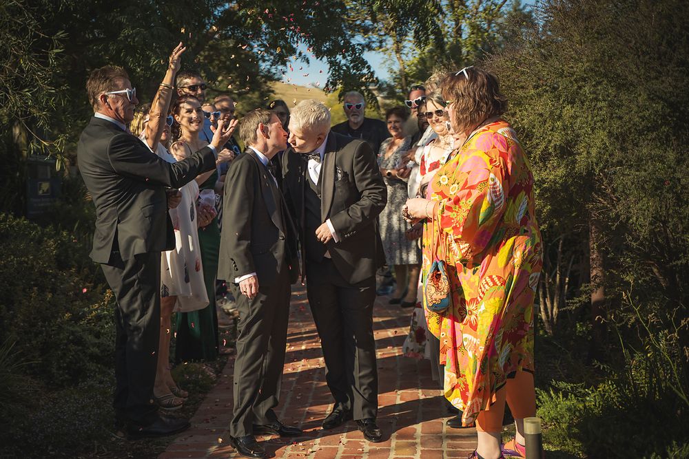 Wedding party gathered outdoors in formal attire with vibrant orange accents against natural foliage backdrop.