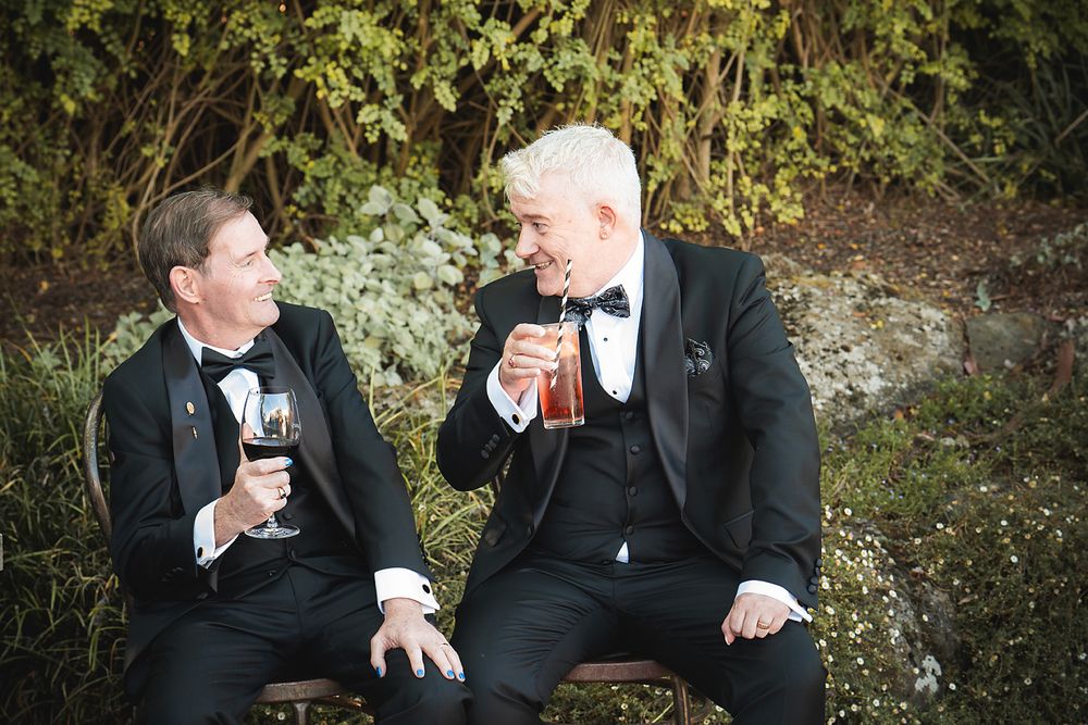 Two people in formal attire sharing drinks and conversation in an outdoor garden setting at dusk.