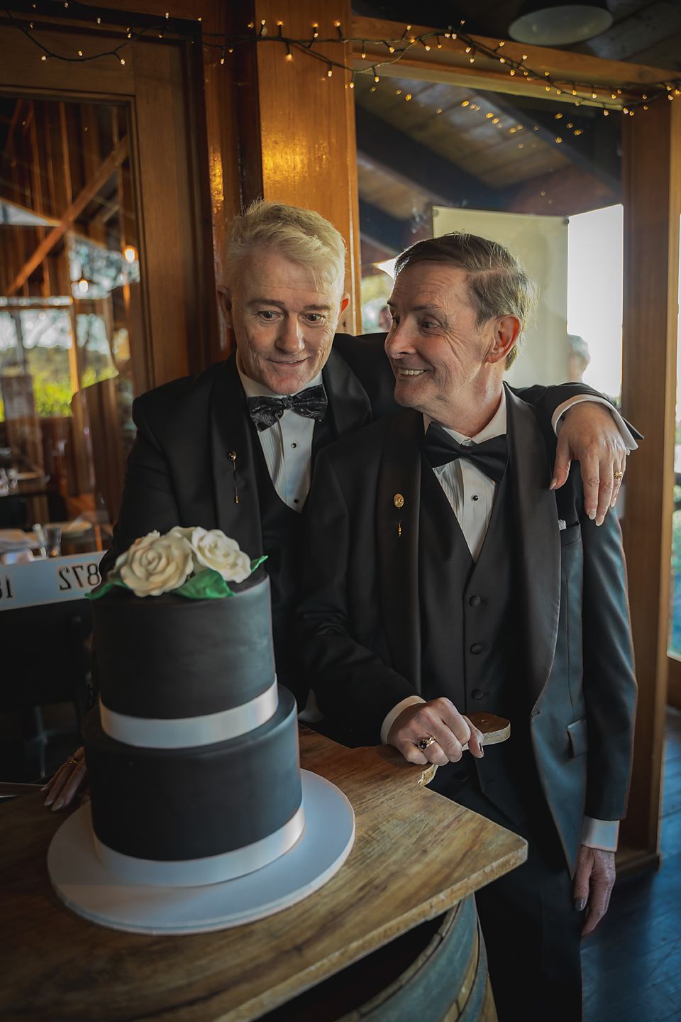 Two people in tuxedos stand together smiling next to a modern three-tier wedding cake with white and navy blue layers.