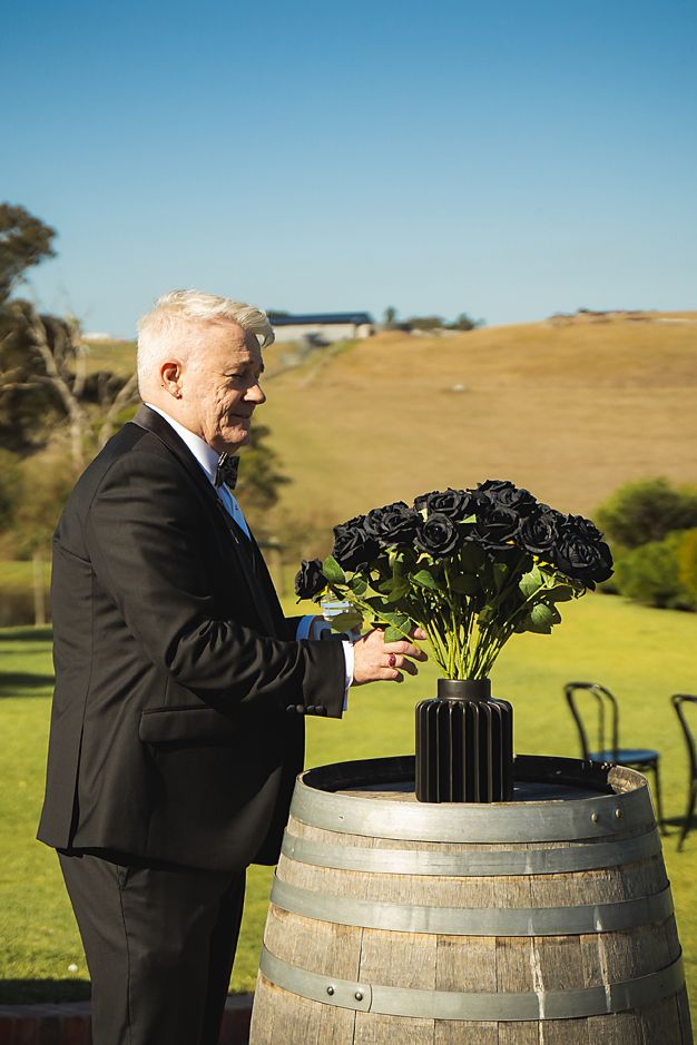 Someone in formal attire arranges black roses on top of a wooden wine barrel in an outdoor setting with rolling hills.