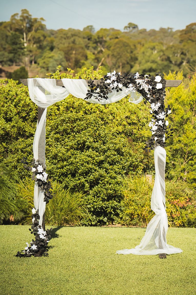 White wedding arch with floral decorations stands on a lush green lawn surrounded by yellow flowering bushes in a garden.