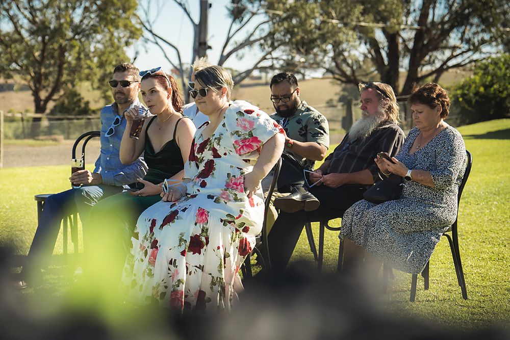 Wedding guests gather outdoors on a sunny day for a casual celebration in a grassy park setting.