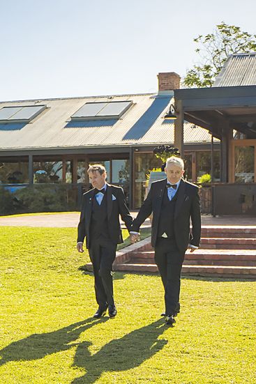 Two people in formal black suits walk together on a sunny lawn in front of a building with skylights.