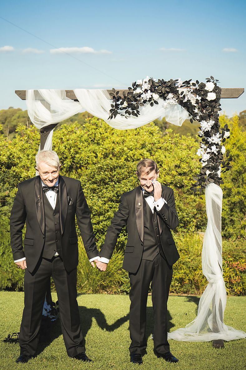 Two people in black tuxedos stand under a white wedding arch decorated with greenery at an outdoor ceremony.