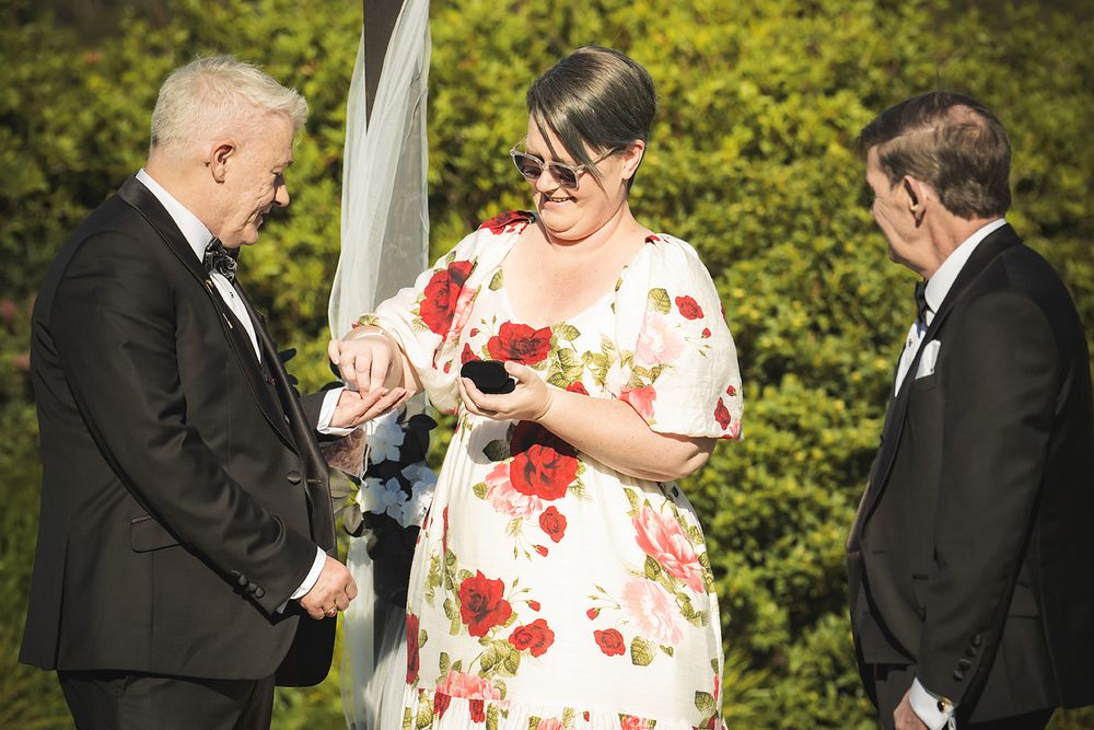 A couple exchanges rings during an outdoor wedding ceremony with floral decor and greenery in the background.