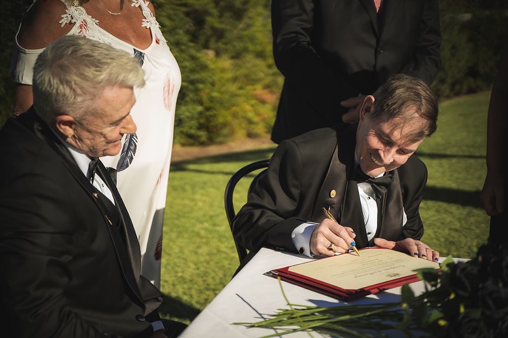 Two people in formal attire sign official documents at an outdoor wedding ceremony.