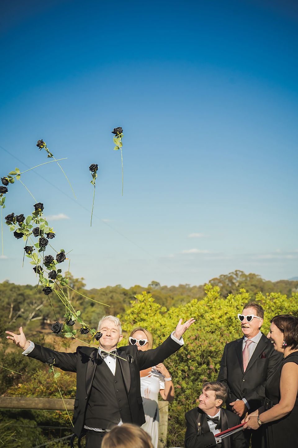 Wedding party members playfully toss flower bouquets into the blue sky during an outdoor celebration.