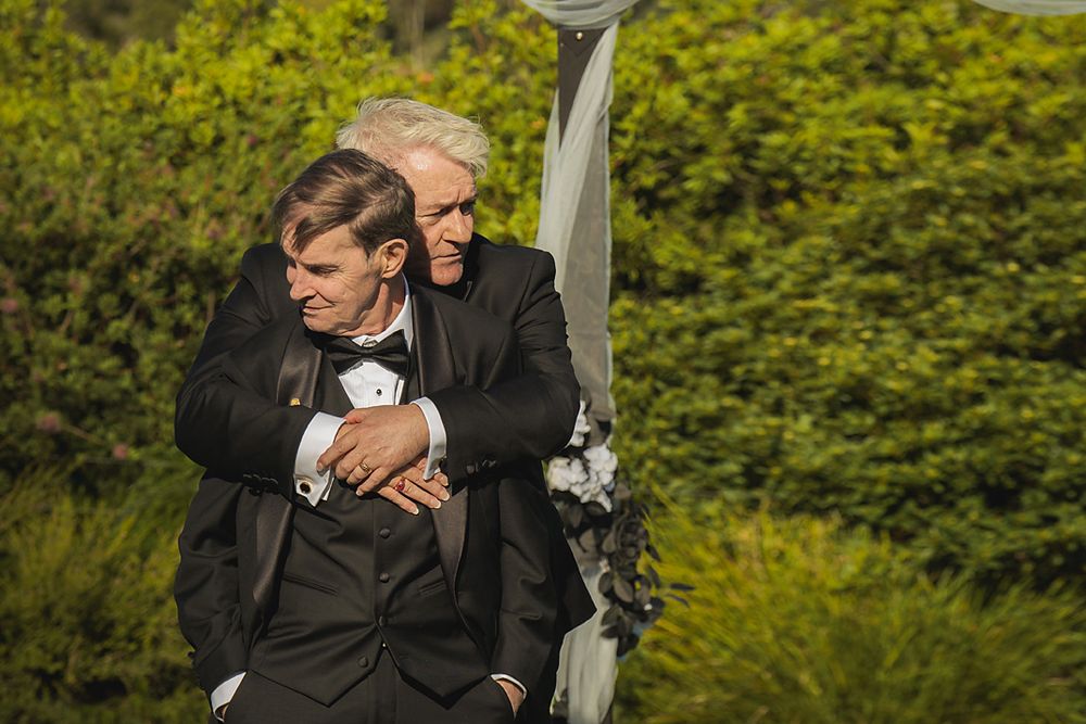 Two people in black suits embrace while seated on a ski lift chair against a green foliage background.
