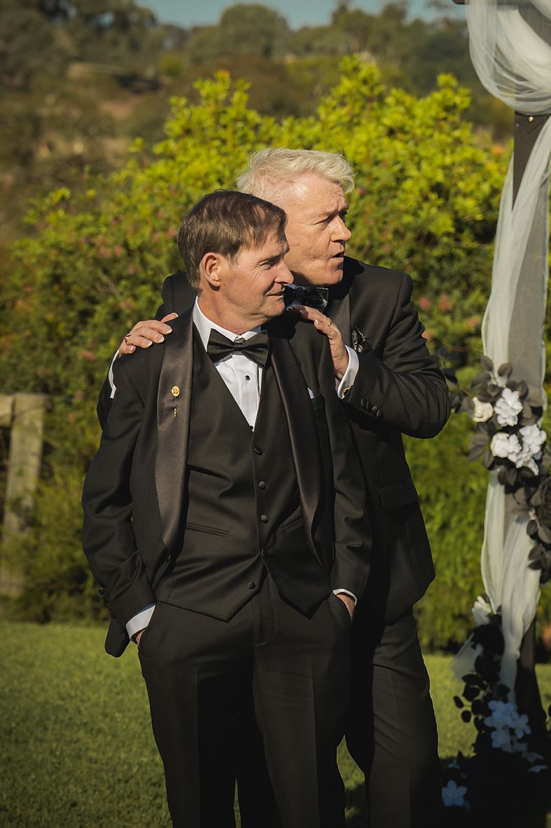 Two men in black tuxedos share a lighthearted moment during an outdoor wedding ceremony.