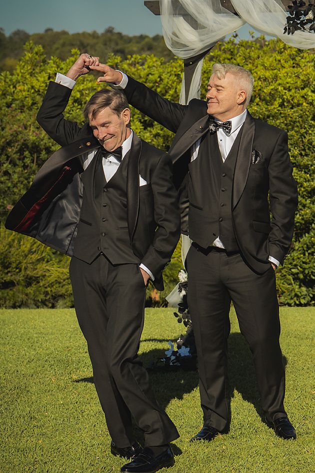 Groomsmen in black tuxedos strike playful poses on a sunny lawn with greenery in the background.