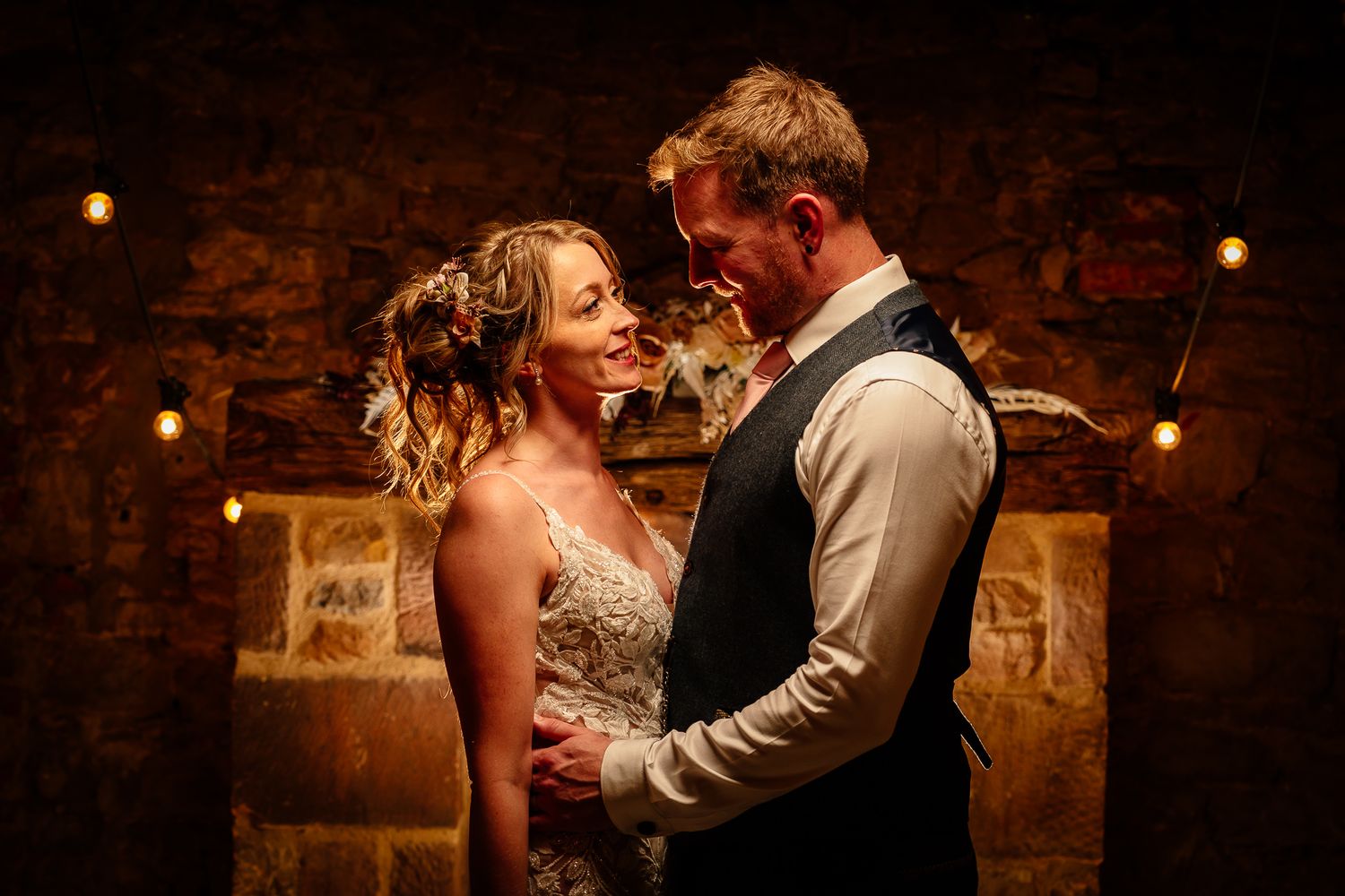 Romantic wedding couple embraces in moody lighting against stone wall backdrop.