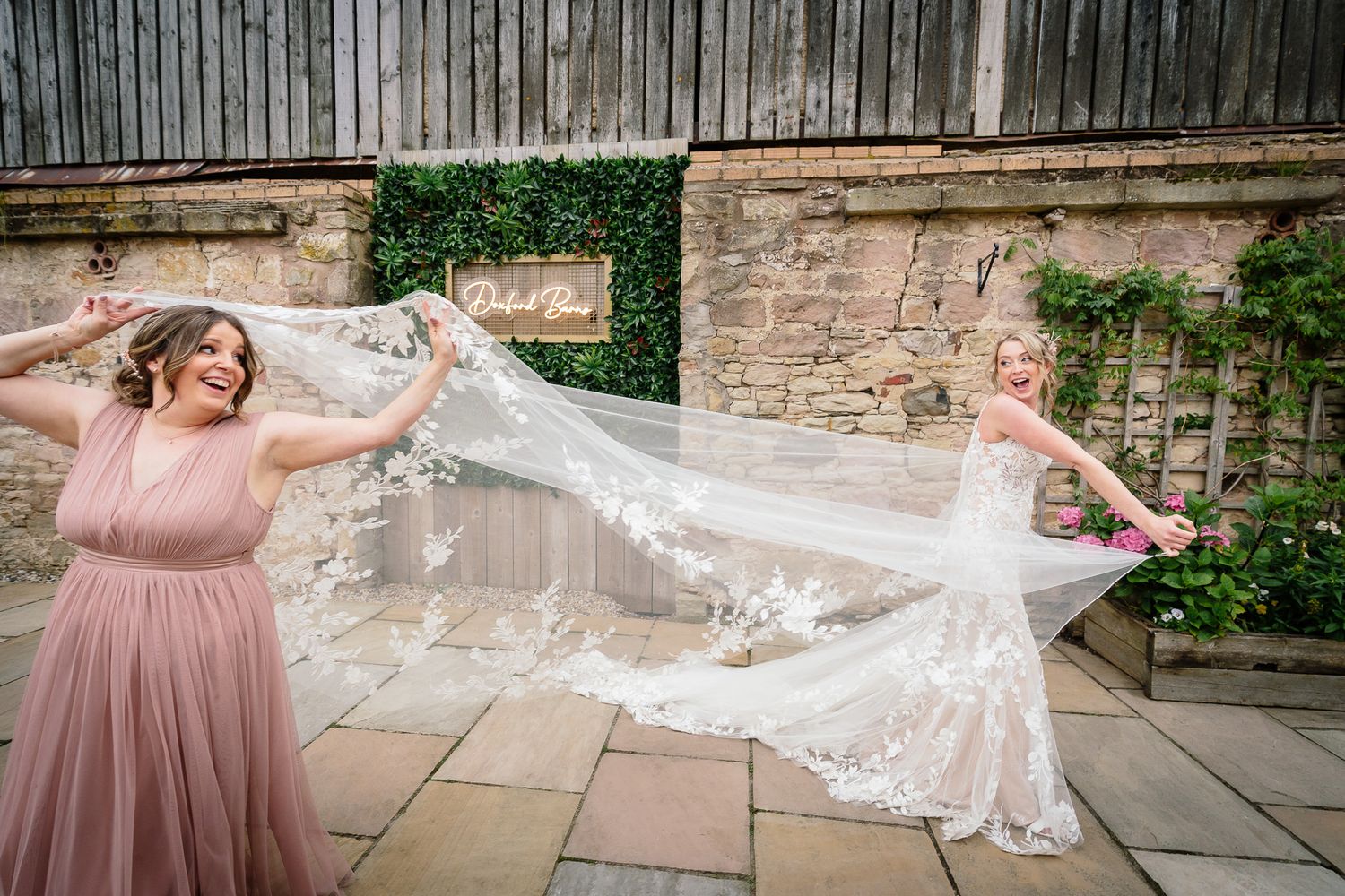 Bride in lace dress with flowing veil against stone wall background.