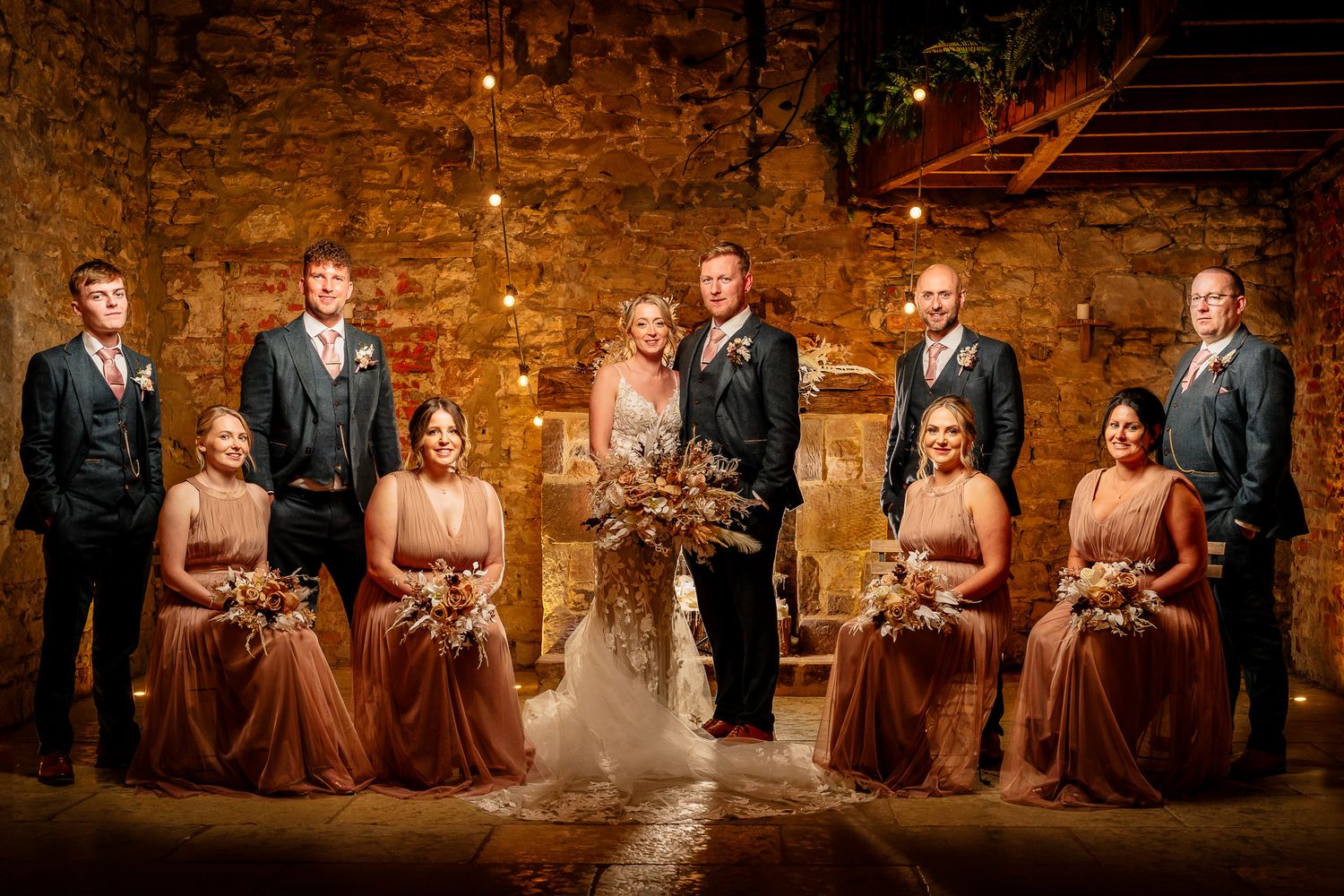 Wedding party group photo with bridesmaids in dusty rose dresses against stone wall.