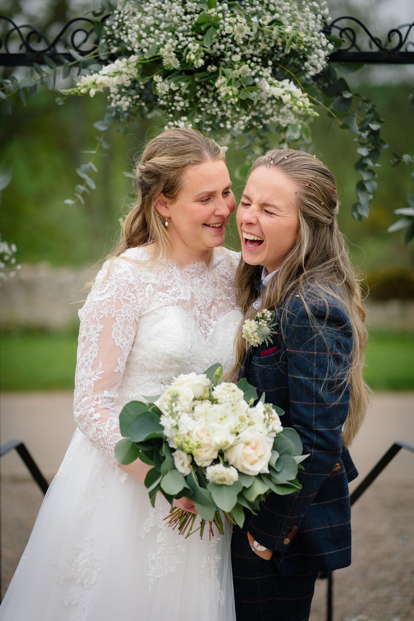 Candid moments under white floral blooms show shared laughter and a beautiful bridal bouquet.
