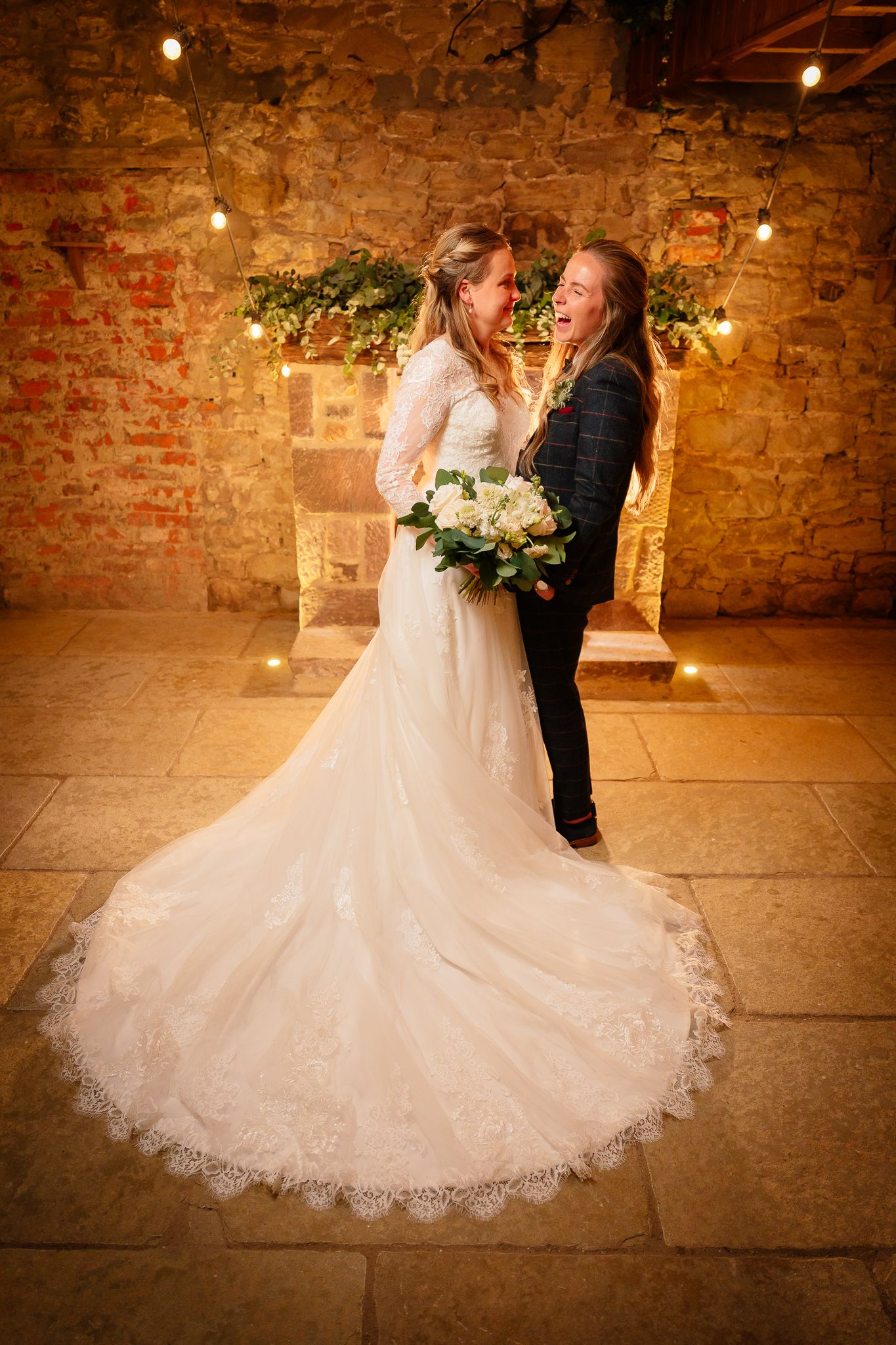 Elegant lace wedding dress train displayed against rustic brick walls with warm ambient lighting.