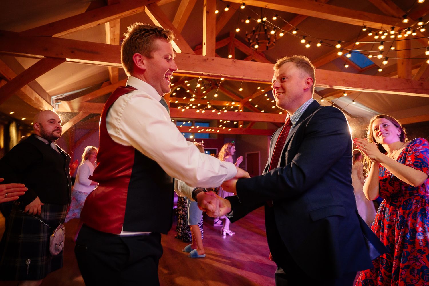 Wedding guests dance energetically under warm string lights at an indoor reception venue.