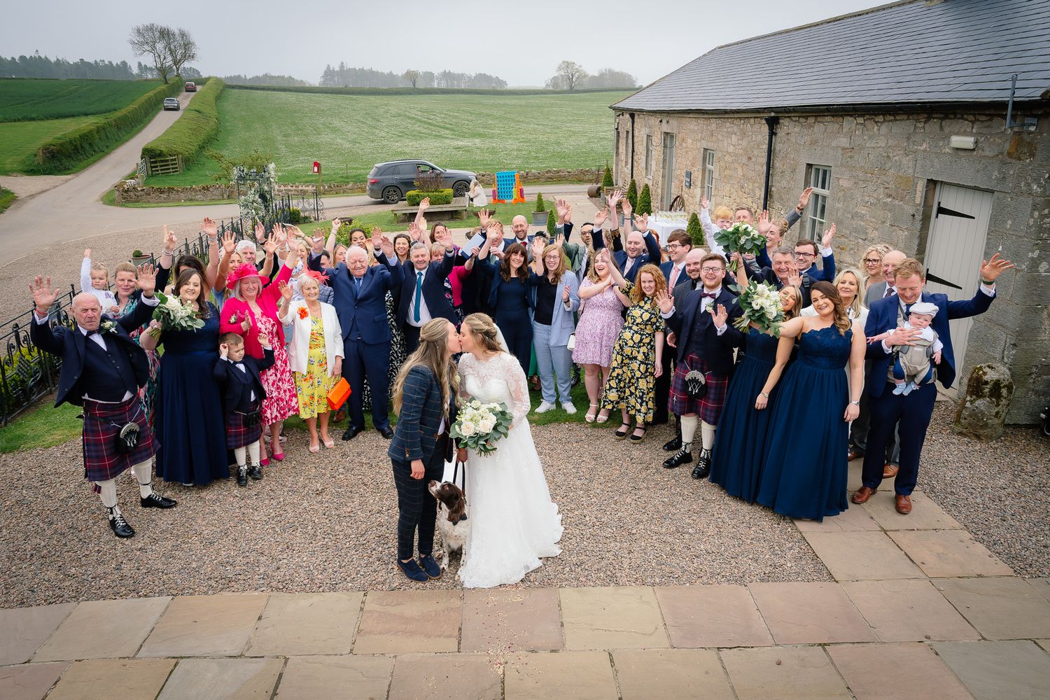 Wedding guests gather outside a rustic stone venue for a group photo celebration.