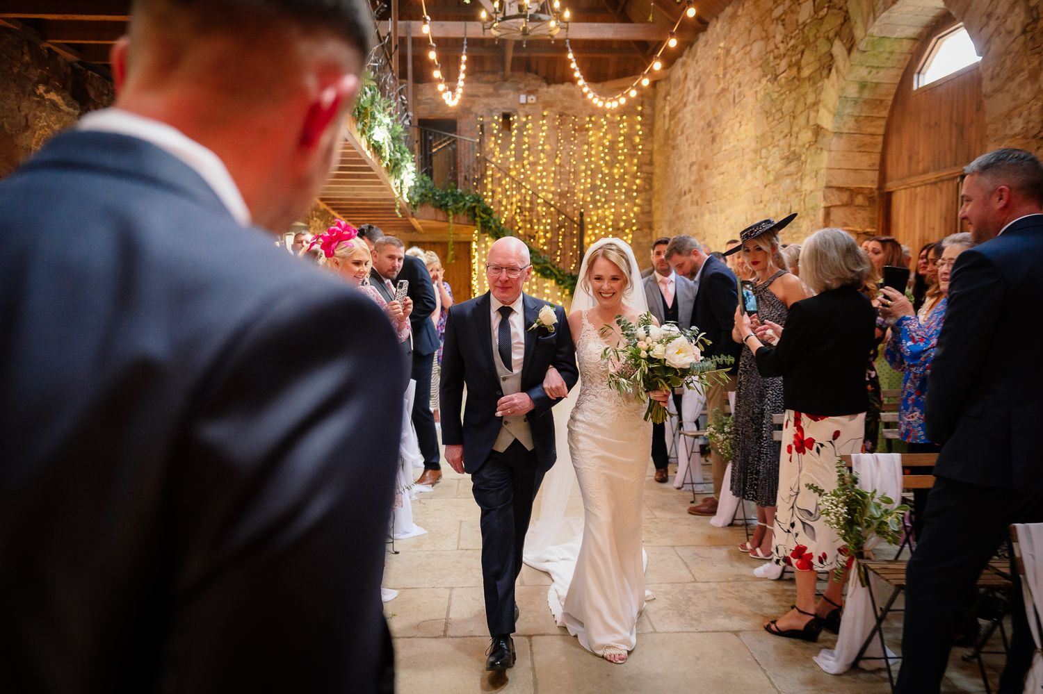 Wedding ceremony in rustic stone barn with string lights and floral decorations.