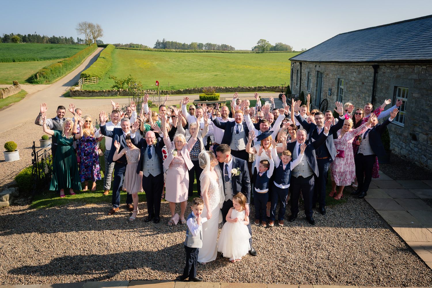 Large group wedding photo taken outside rustic barn venue in countryside setting.