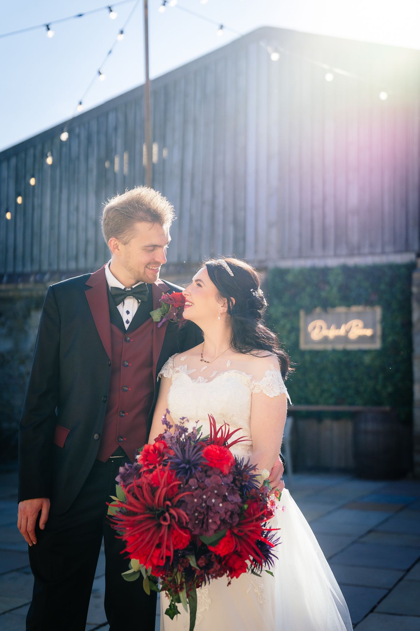 Wedding couple poses with vibrant red bouquet against industrial backdrop. Doxford Barns