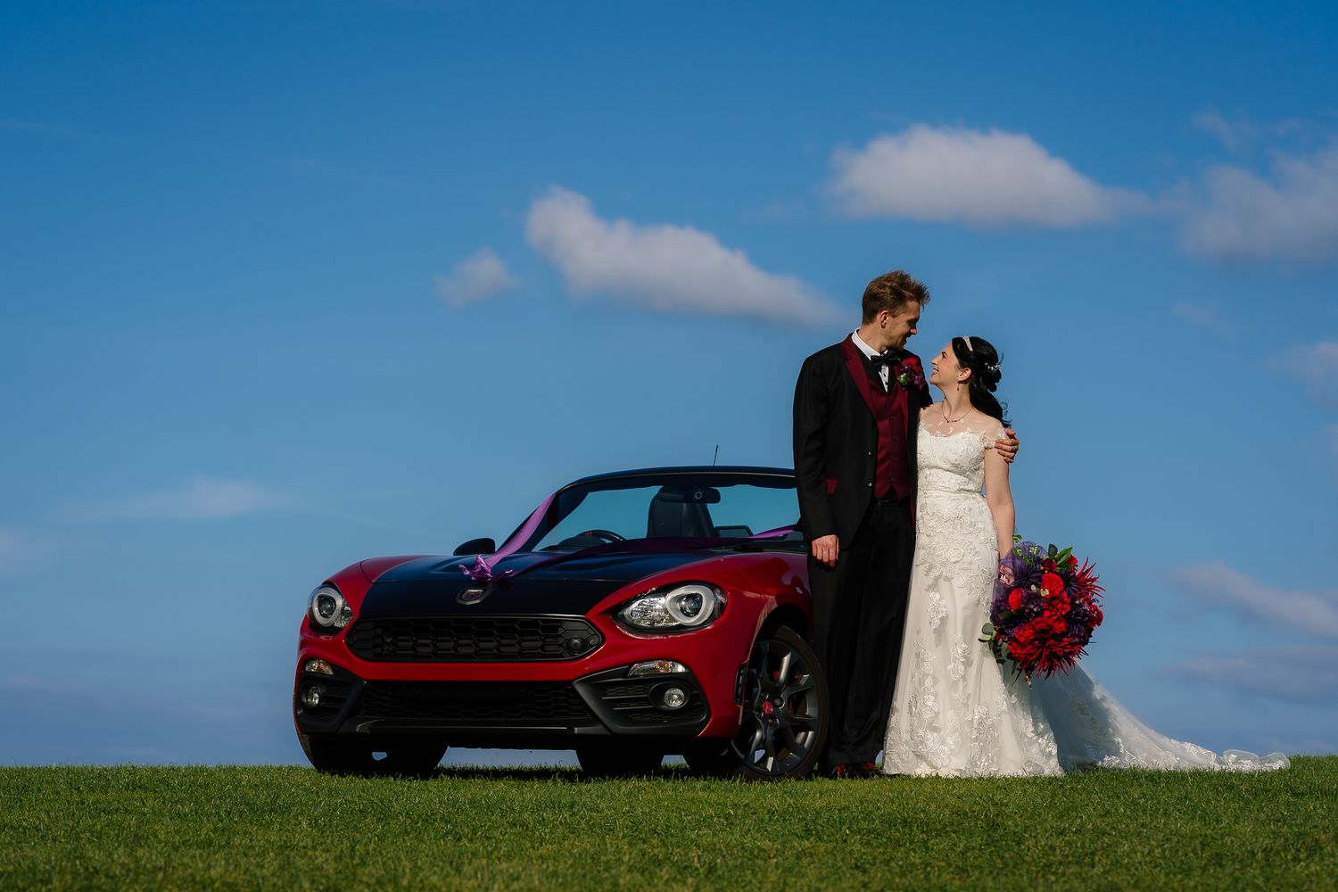 Red sports car parked on a grassy hill against blue sky with white clouds during a wedding photoshoot. Doxford Barns