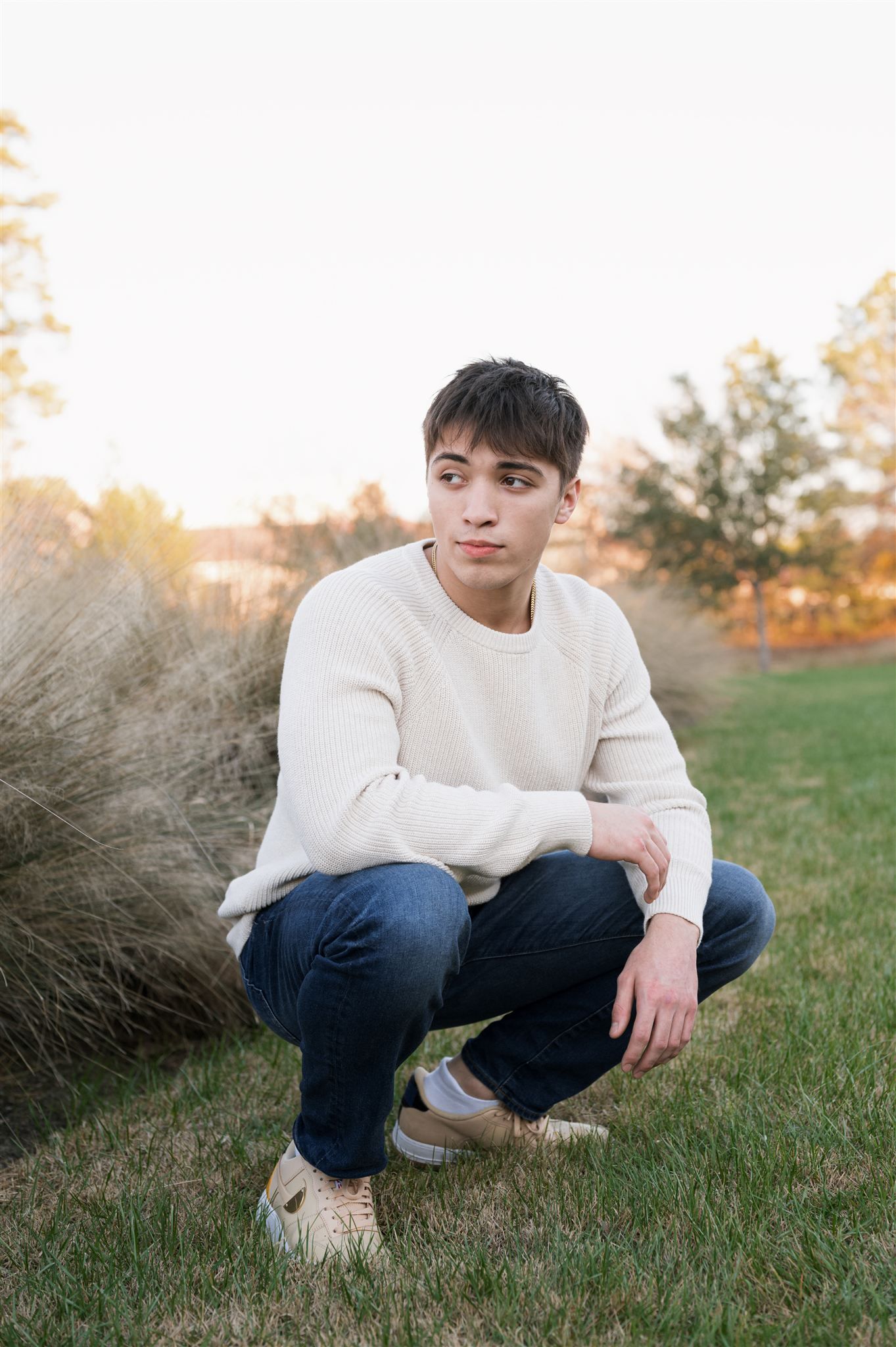 Person in white sweater and jeans crouches on grass during golden hour with autumn foliage in background.