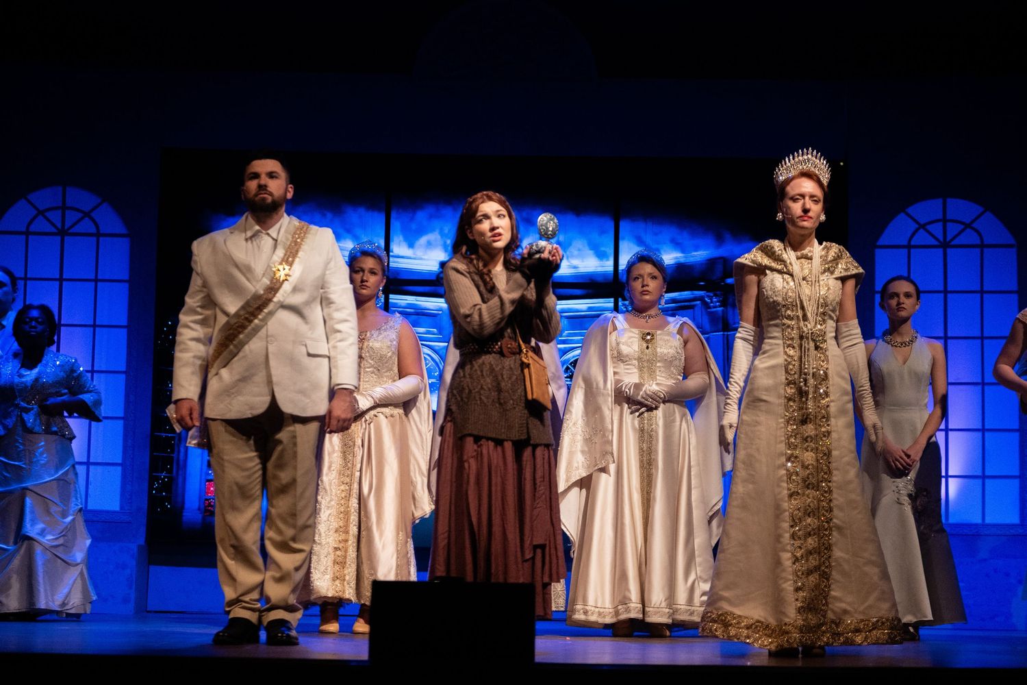 Theater performers in white and beige costumes stand on stage with blue lighting and arched windows in the background.
