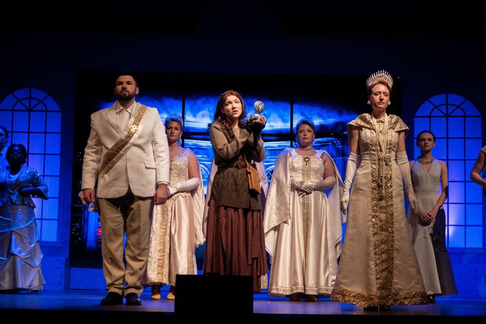 Theater performers in white and beige costumes stand on stage with blue lighting and arched windows in the background.