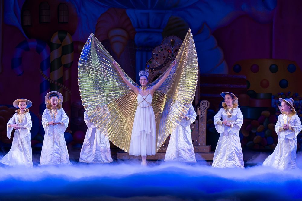 Performers in white angel costumes with illuminated wings dance on a dark blue-lit stage during a theatrical production.