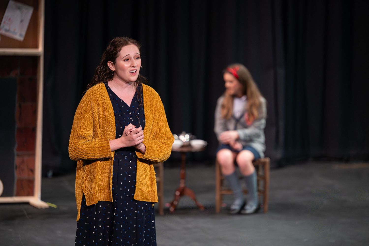 Two women performing on stage, one in a yellow cardigan speaking, another seated in background during a theater production.