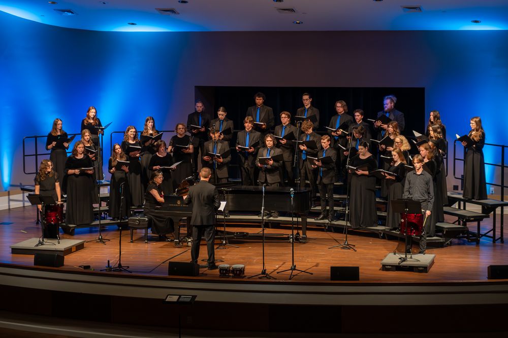 A musical performance on stage with blue lighting and multiple performers arranged in a choral formation.