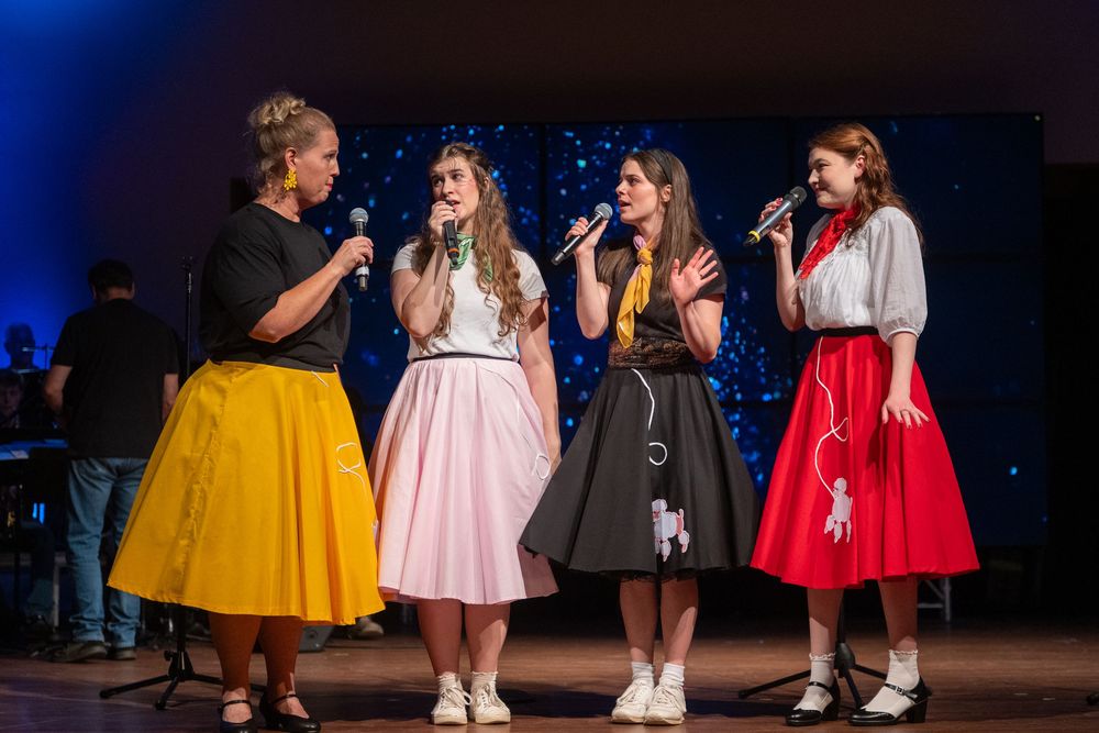 Four performers in vintage 1950s style circle skirts and blouses sing together on a dark stage during a show.