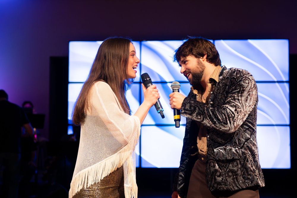 Two performers singing together passionately on stage with dramatic lighting and video screens in the background.
