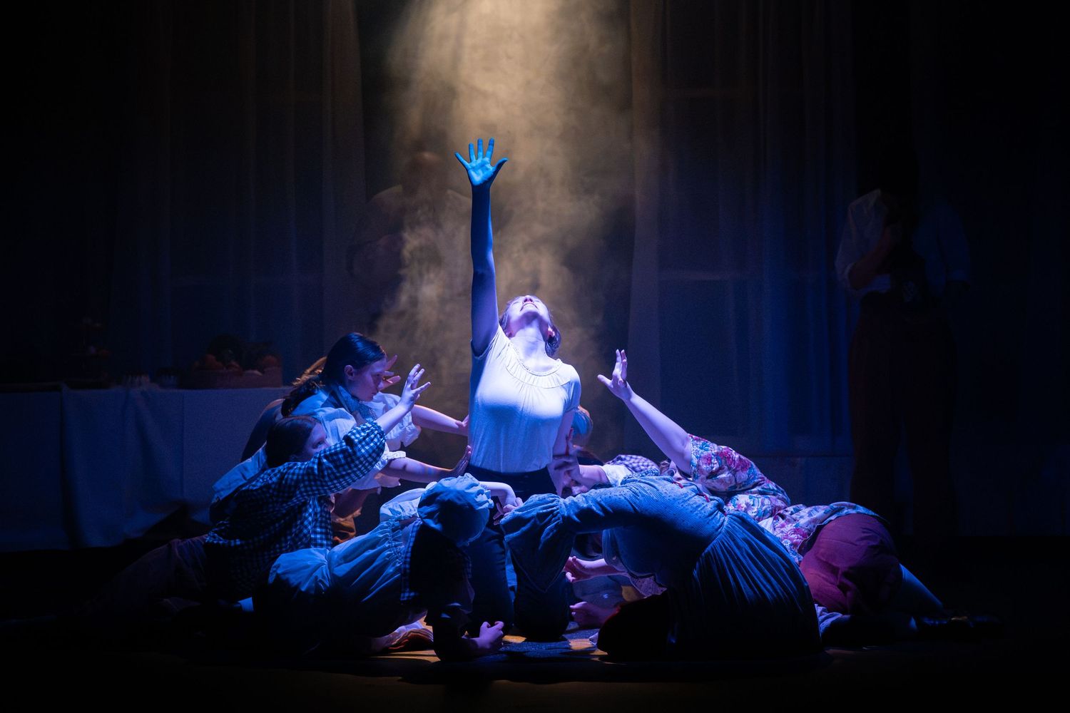 Dramatic stage performance with dancers in blue light, one performer reaching upward with glowing hand.