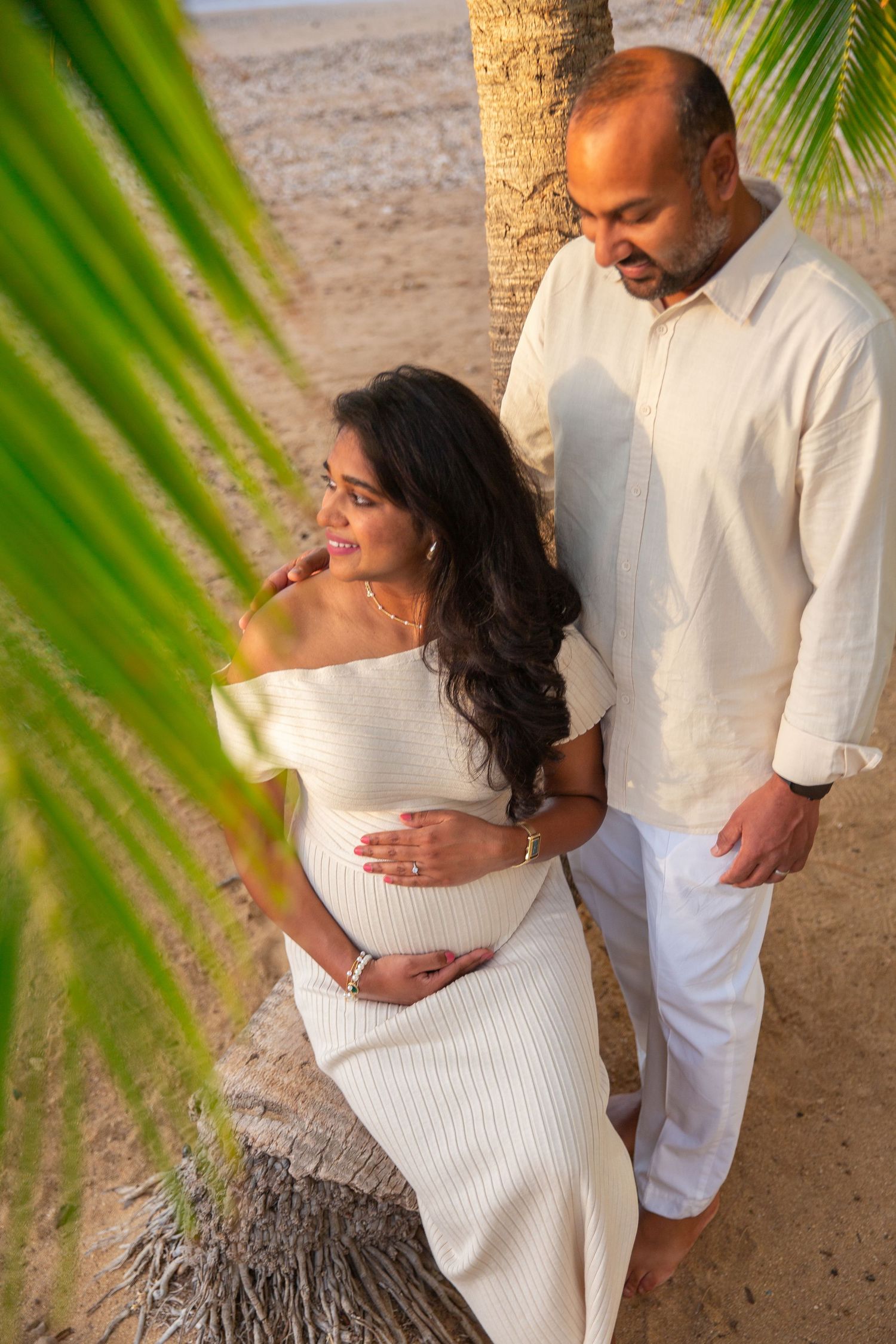 A couple in white outfits shares intimate moments on a tropical beach with palm fronds framing the romantic scene at Waialae Beach.