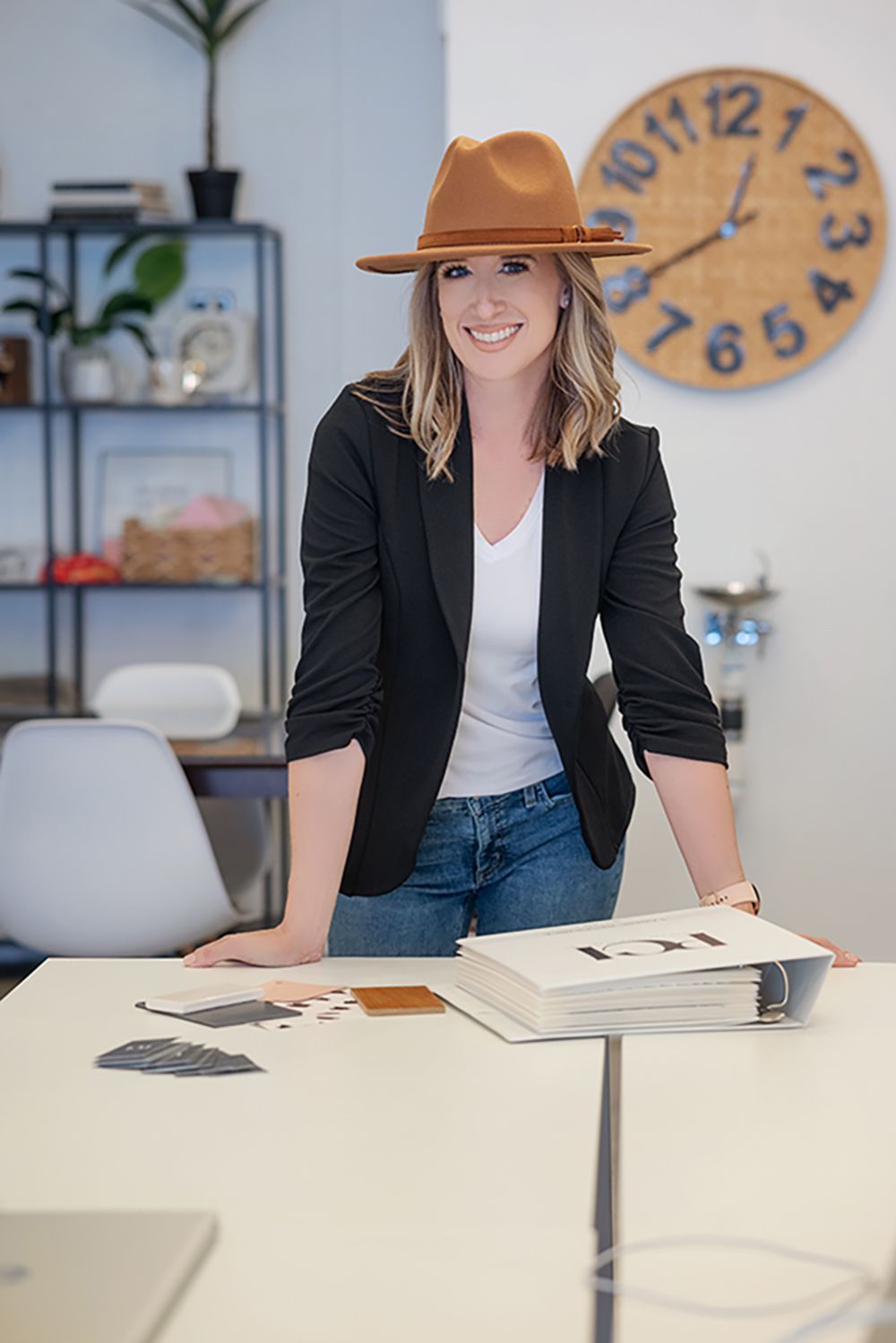 Professional in a black blazer and baseball cap works at a modern desk with decorative wall clock in background.