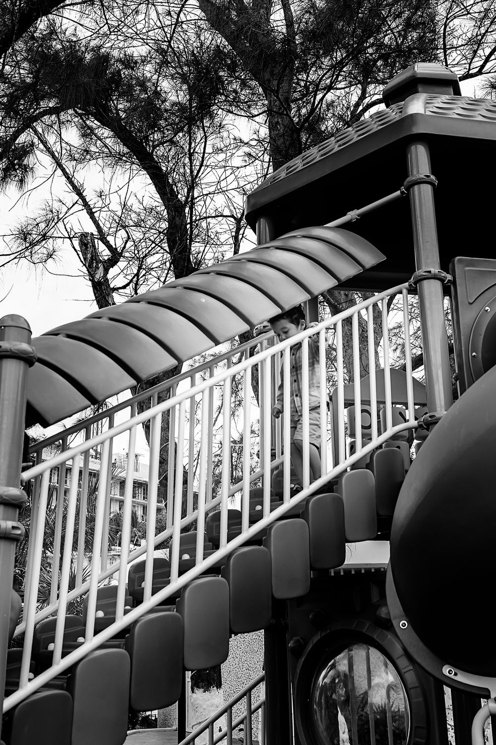 Black and white view of a curved playground slide with metal railings against a backdrop of trees.